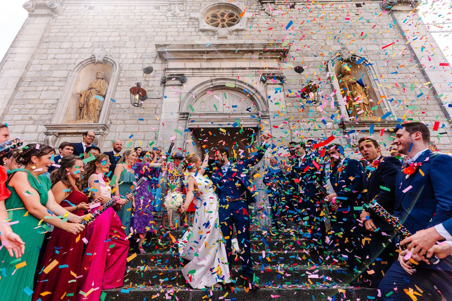Wedding guests celebrate outside a historic stone church as colorful confetti rains down during the joyful ceremony exit.