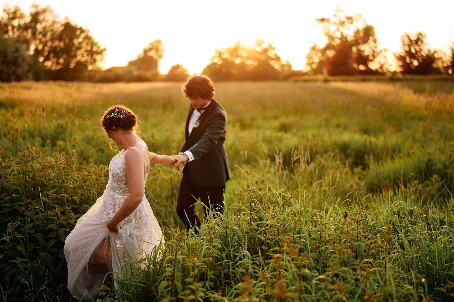 A couple walks hand in hand through a sunlit meadow during golden hour while wearing formal wedding attire.