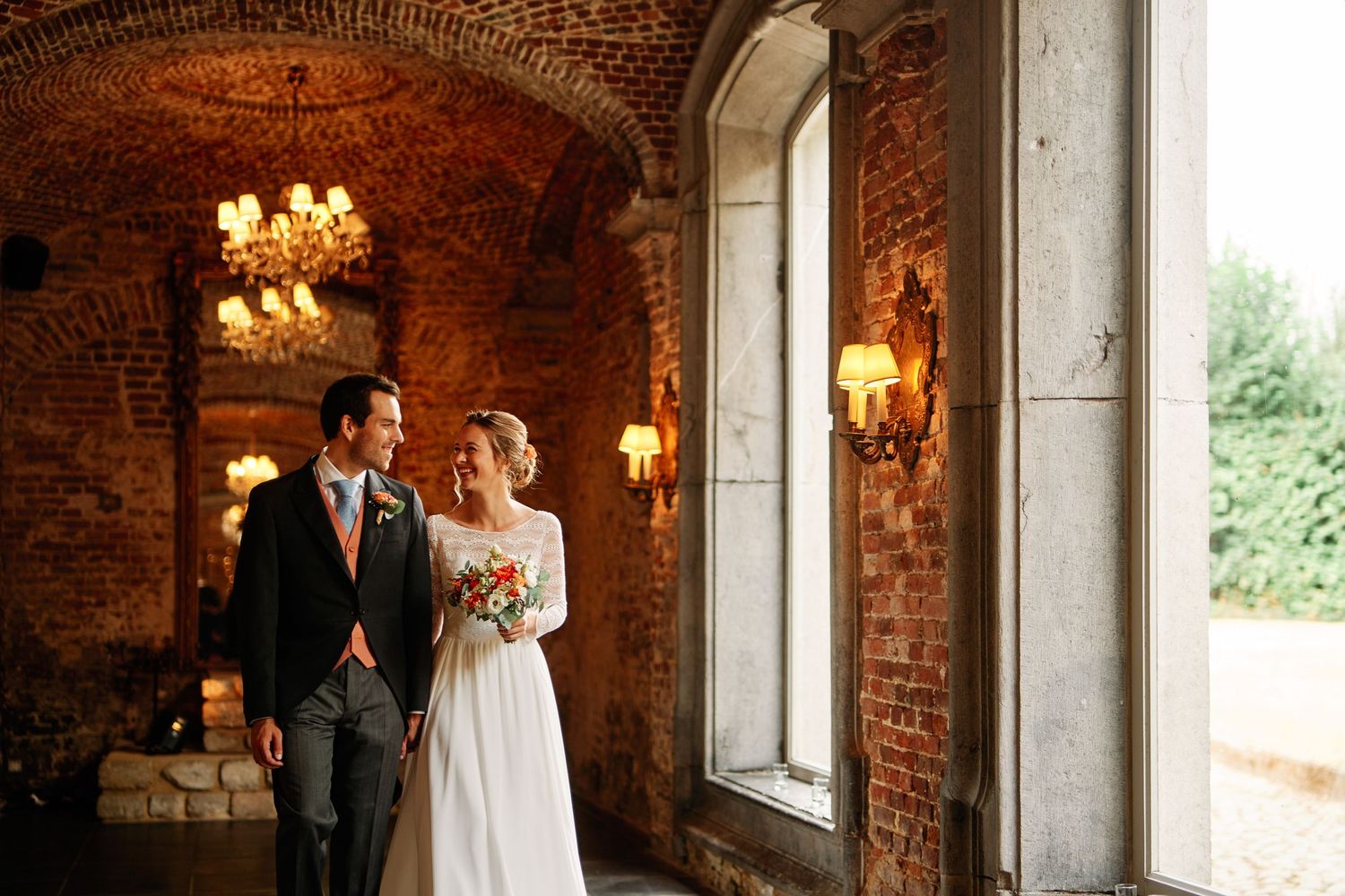 Romantic couple stands together in rustic brick venue with elegant chandeliers and natural window lighting.