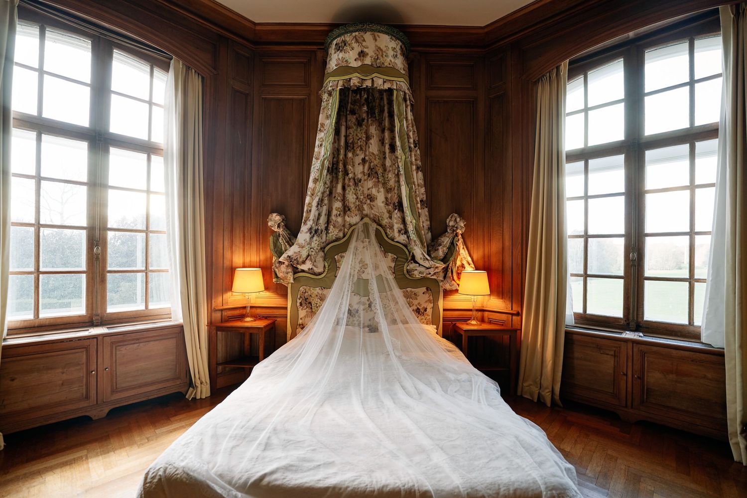 A panoramic view of an elegant bedroom with wood-paneled walls, tall windows, and a canopy bed with flowing white drapes.