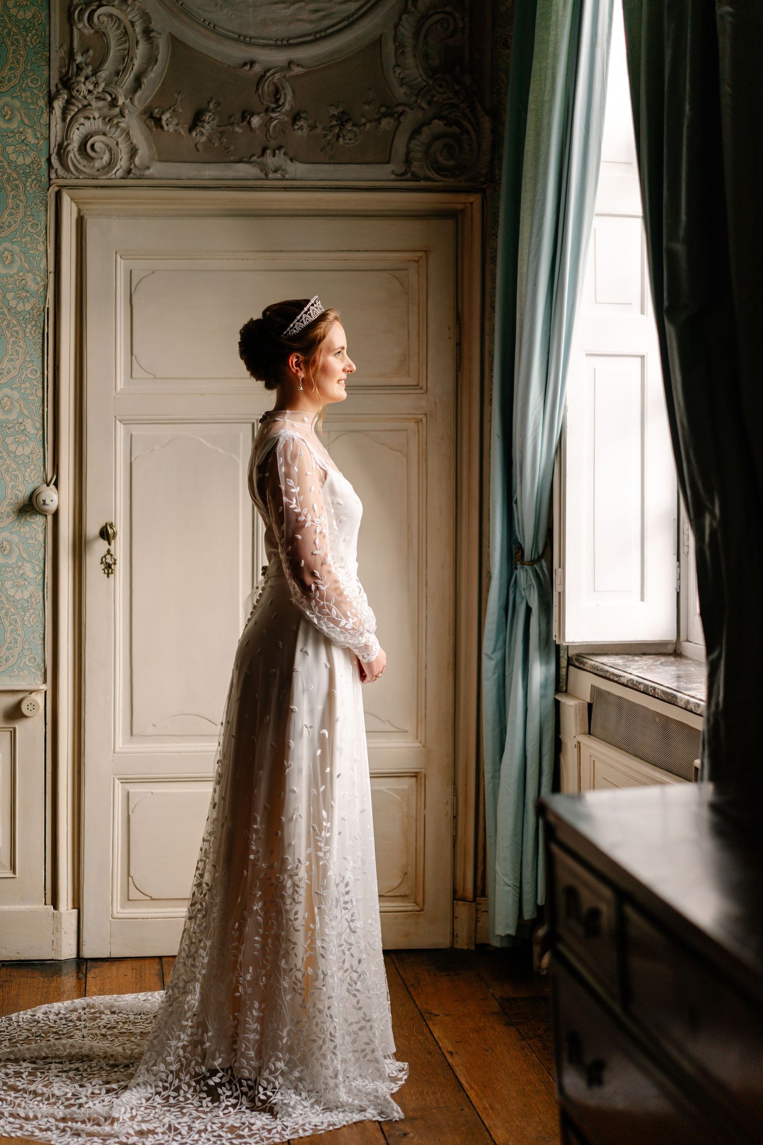 A bride in a vintage-style lace wedding gown stands elegantly by a window in an ornate room with turquoise curtains.
