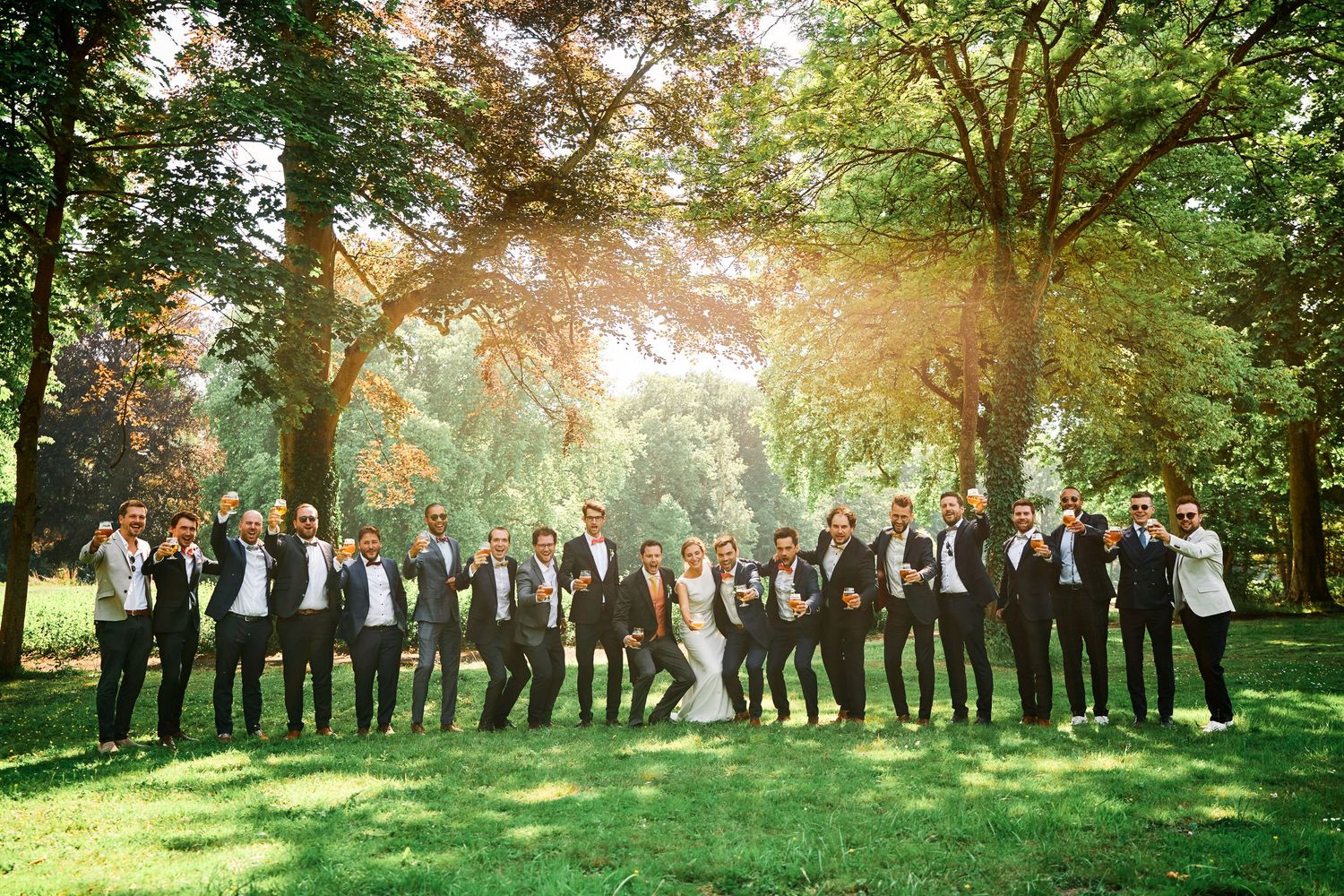 A large wedding party stands in a line outdoors under trees with golden sunlight filtering through the branches.