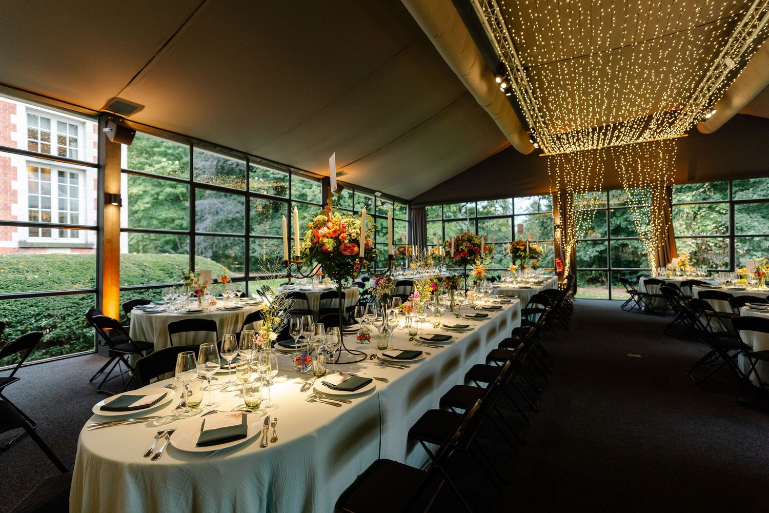 Elegant wedding reception venue with long white tables, string lights, and floor-to-ceiling windows overlooking lush greenery.