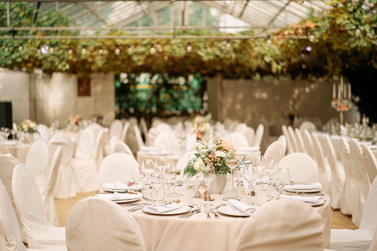 Elegant wedding reception tables set with white linens and floral centerpieces in a glass-enclosed venue with natural lighting.