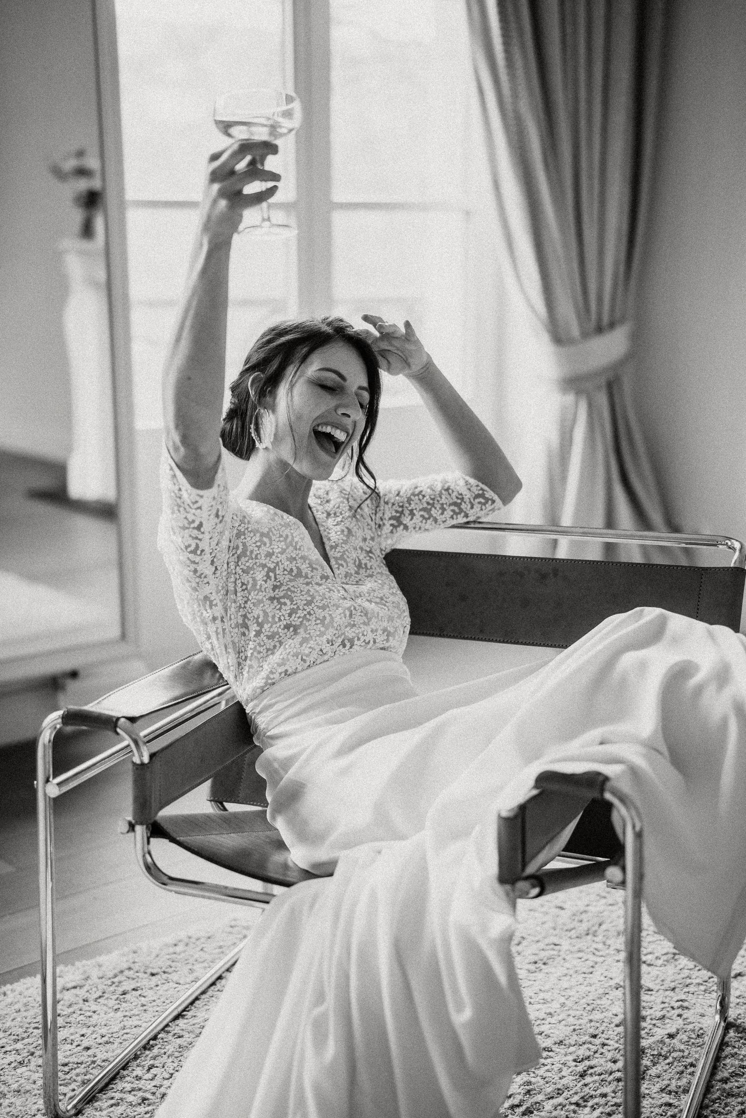 A bride in a white lace gown poses gracefully on a chaise lounge in a bright, elegant room.