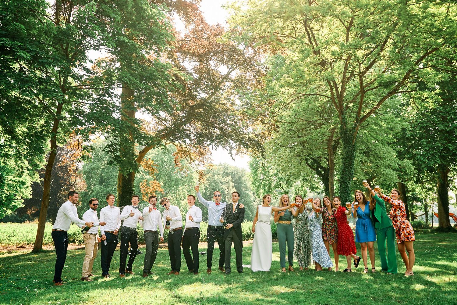 Wedding party stands in a line under lush green trees during an outdoor summer celebration.