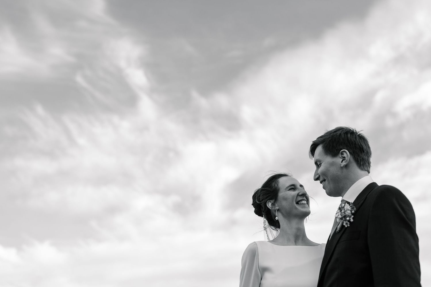 A black and white photo of a couple sharing a joyful moment against a dramatic cloudy sky.