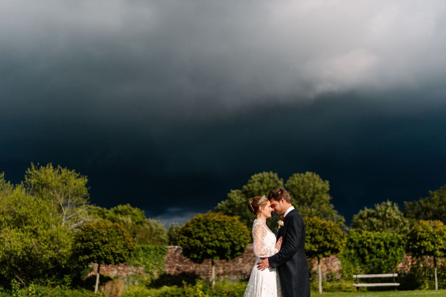 A wedding couple stands in a dramatic landscape with dark storm clouds looming overhead and green trees in the background.