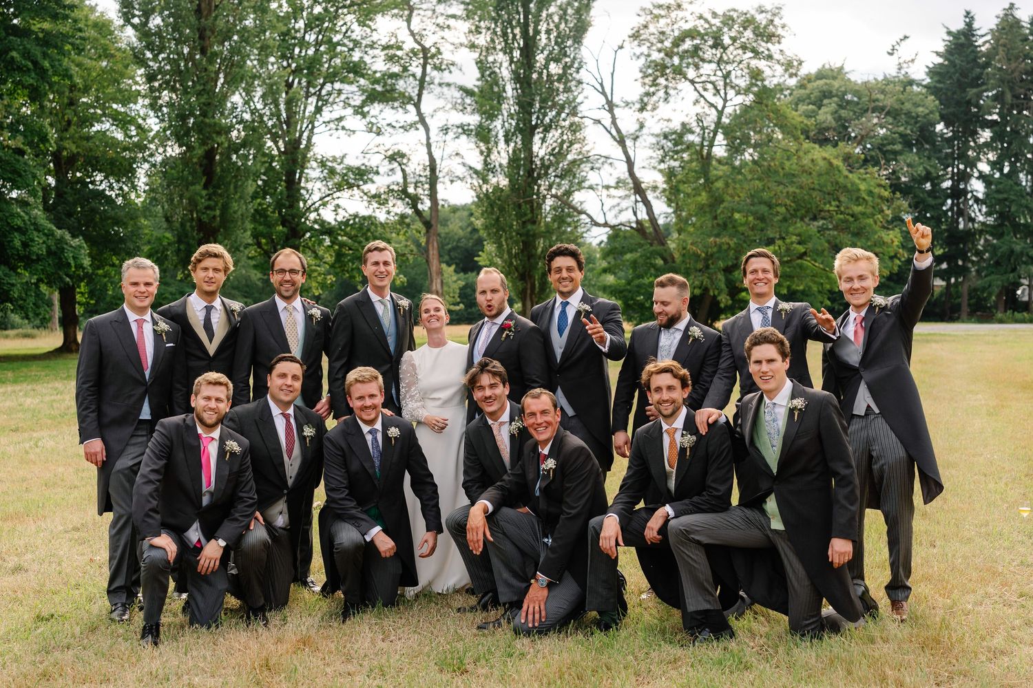 Groomsmen in dark suits strike playful poses together on grassy field during outdoor wedding photo session.