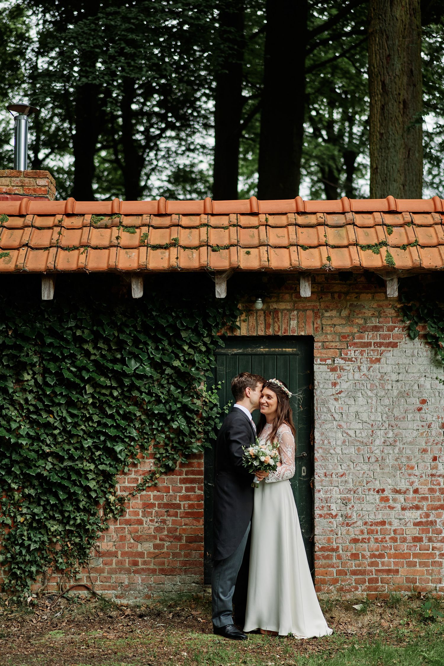 A couple embraces by an ivy-covered brick wall with terracotta roof tiles and tall trees in the background.