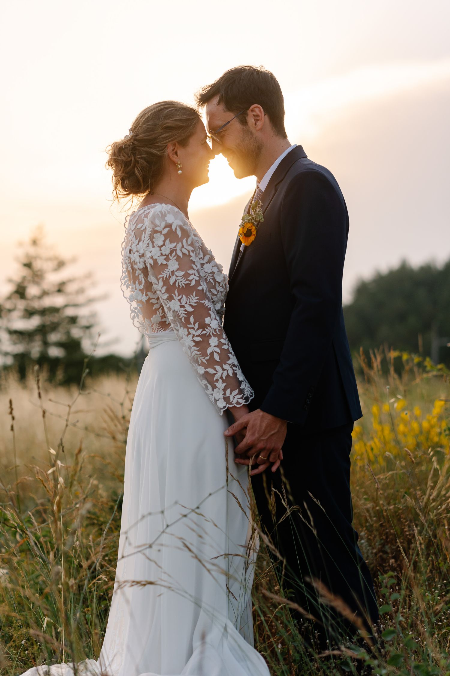 A romantic couple in wedding attire embraces in a golden field at sunset during their outdoor photoshoot.
