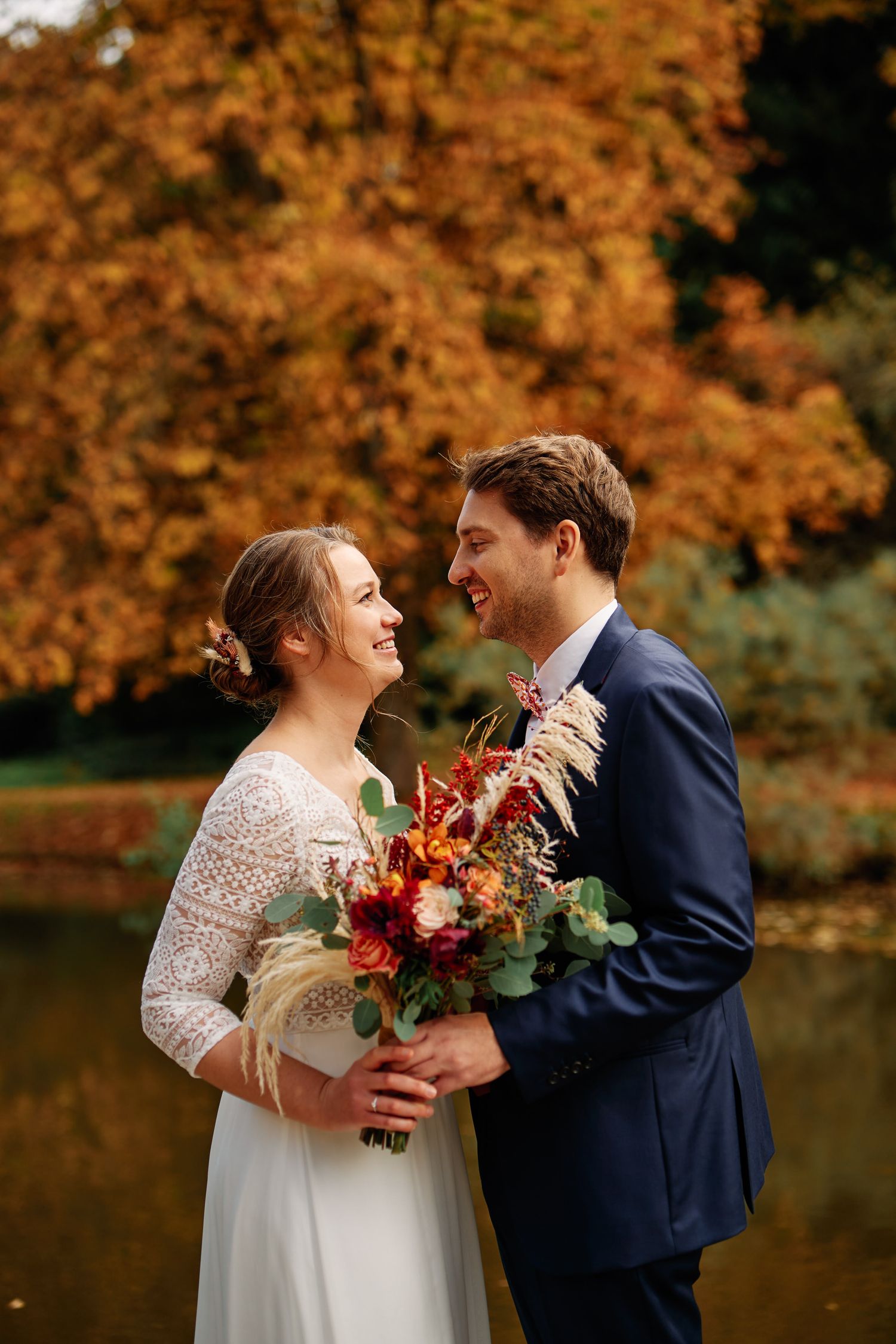 Newlywed couple shares romantic moments in autumn setting with orange foliage and lake backdrop.