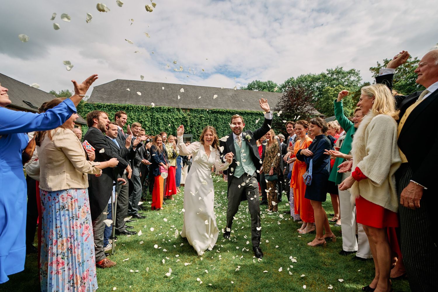 Wedding guests throw flower petals in celebration as a couple walks past during an outdoor ceremony.