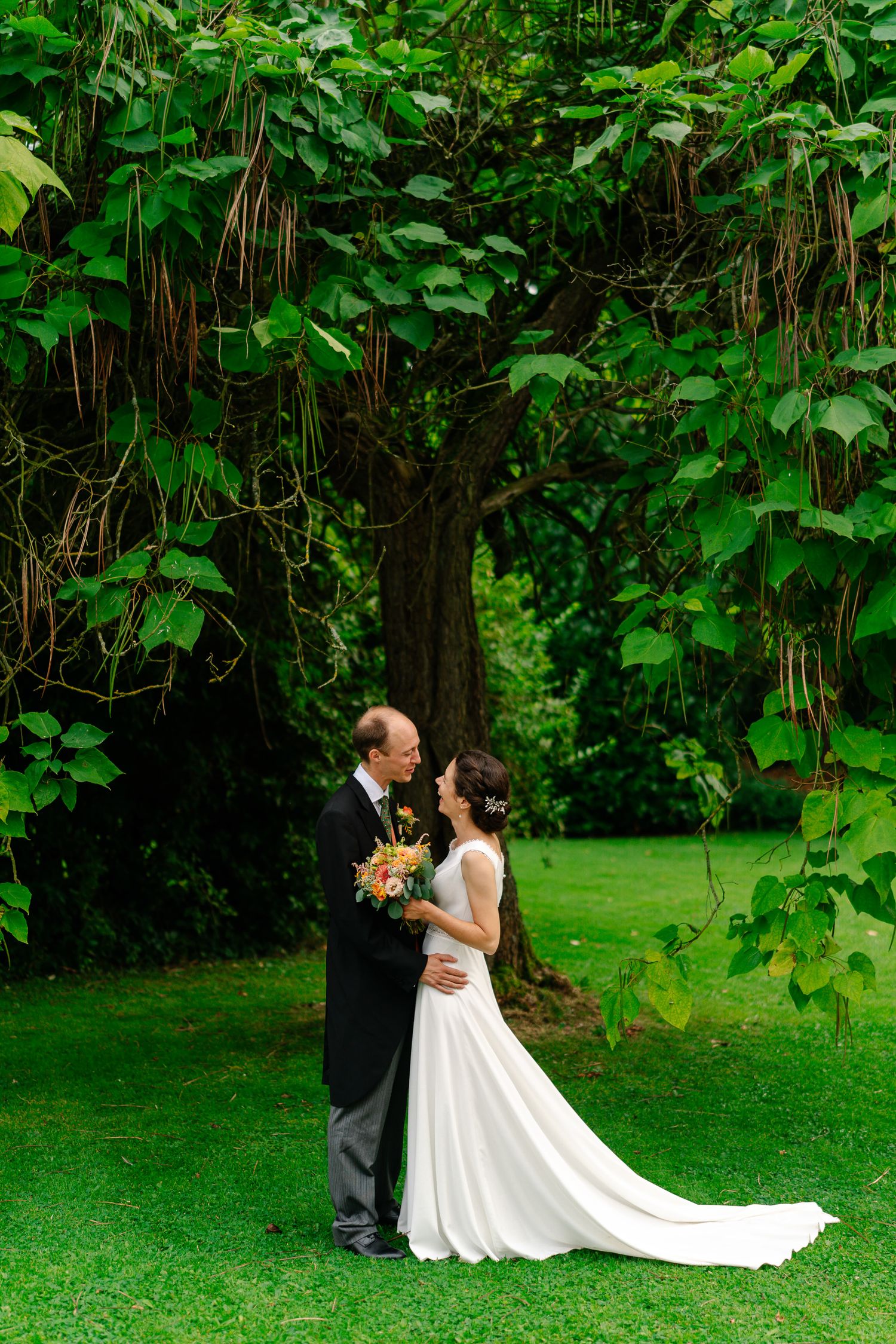 A couple shares a romantic moment in a lush garden setting with trailing white wedding dress and green foliage backdrop.