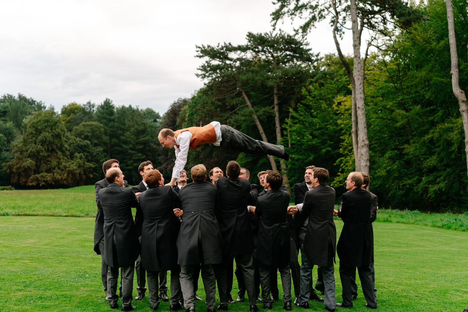 Wedding party in black suits stands in line on grassy field throwing a basketball into the air in sequence.