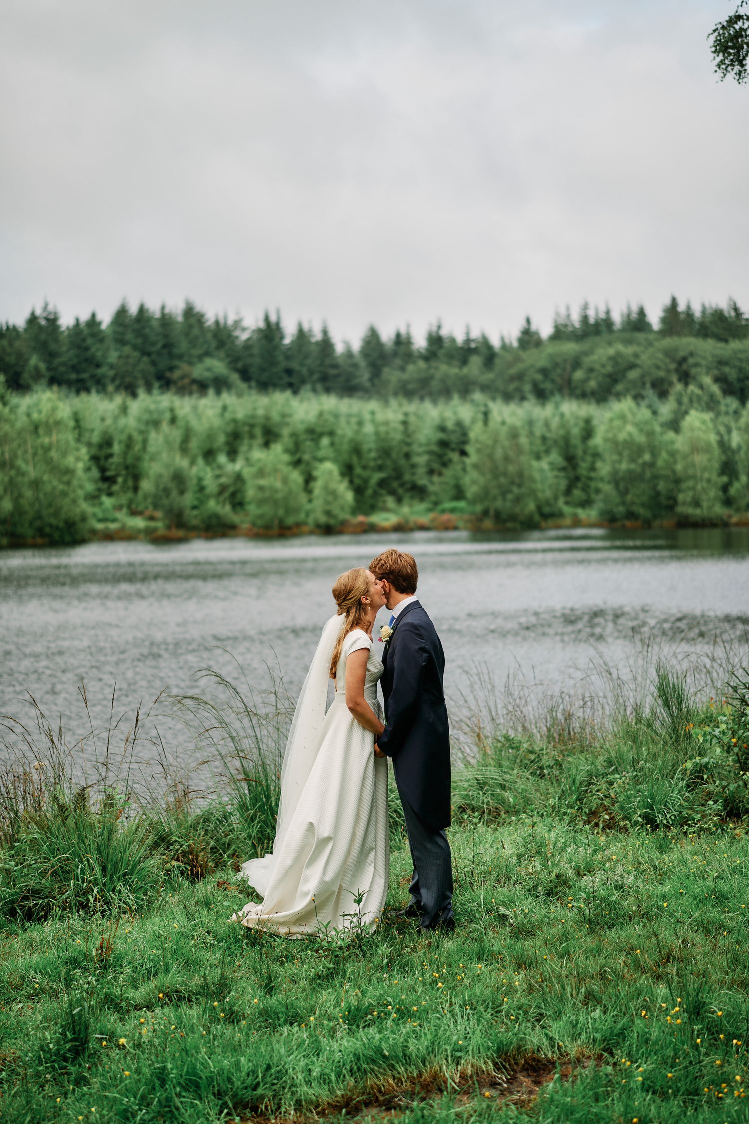 A couple shares a romantic moment by a serene lake surrounded by evergreen forest on their wedding day.