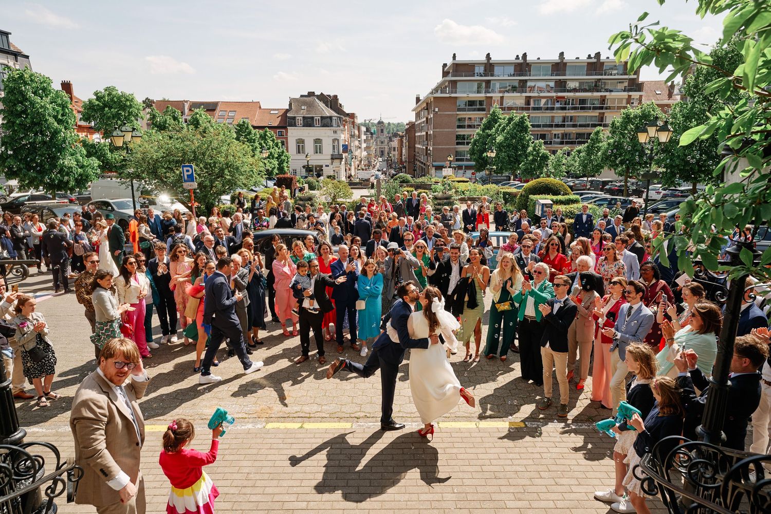 A large crowd of people dancing outdoors at a summer festival with trees and buildings in the background.