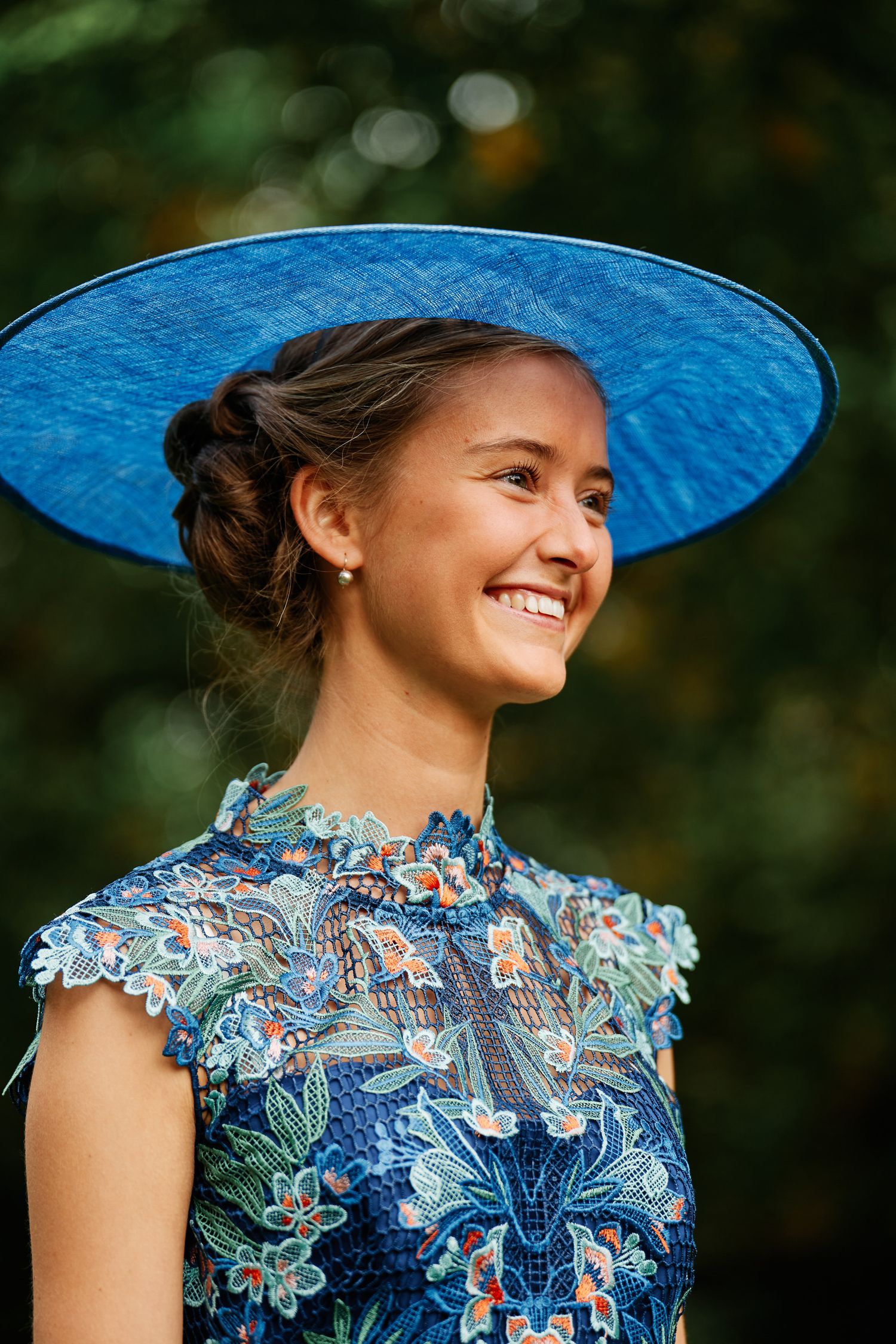 A person wearing a blue wide-brimmed hat and floral dress smiles outdoors against a blurred natural background.
