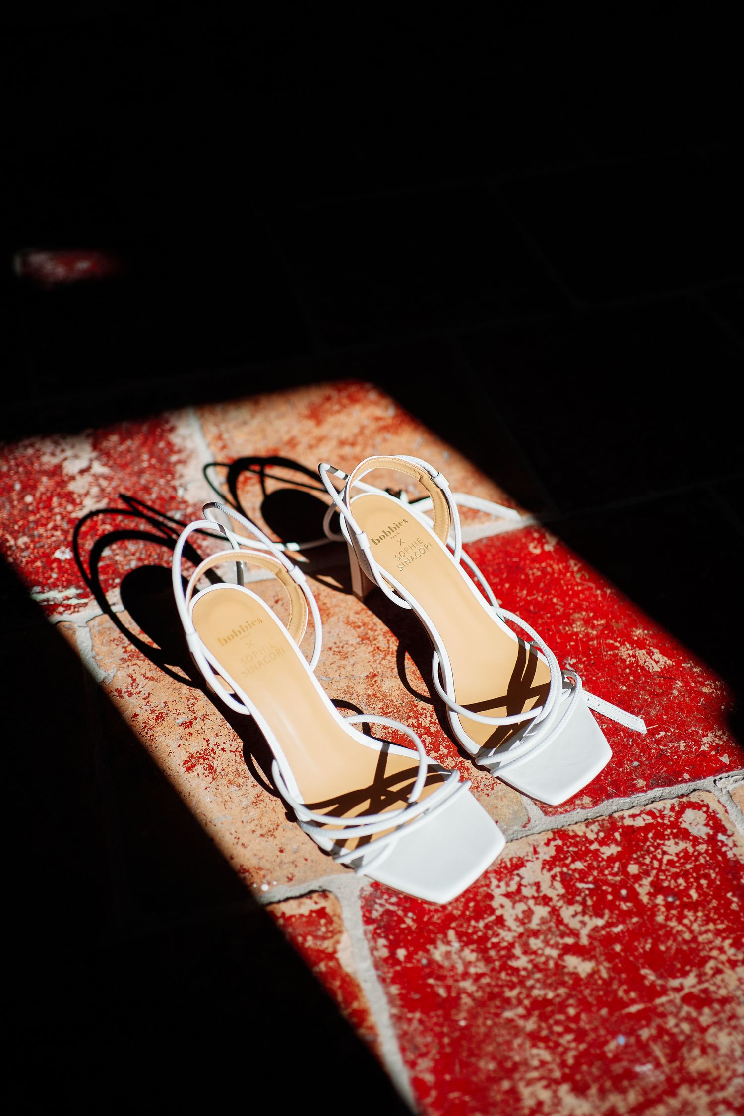 Elegant white strappy sandals with square toes positioned on a textured red surface in dramatic lighting.