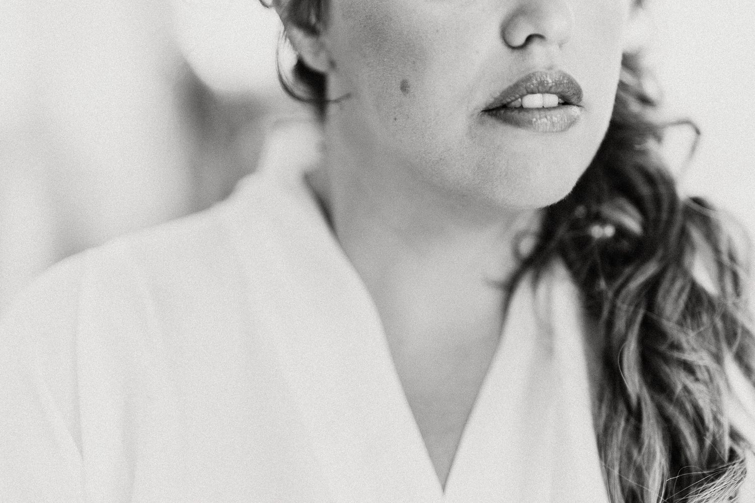Black and white close-up portrait showing lips and curled hair against soft background, wearing white robe.