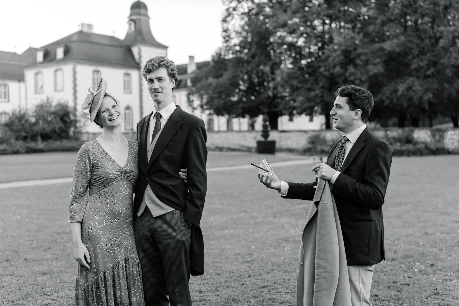 Three people in formal attire share a lighthearted moment on a lawn with a grand house in the background.