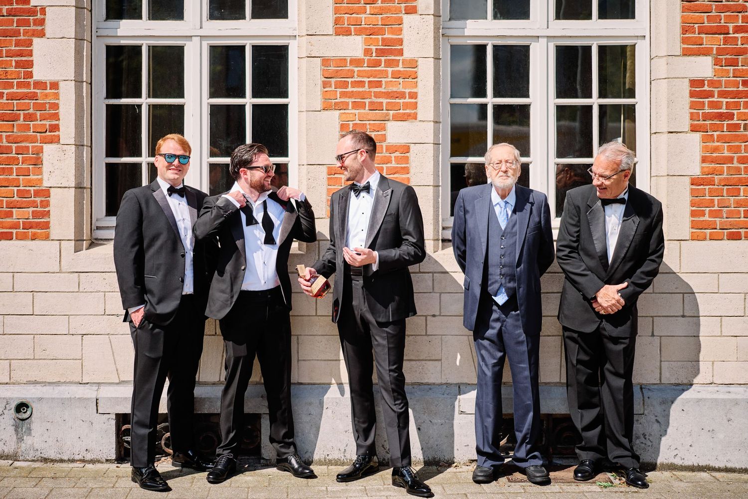 Five men in formal black tuxedos and bow ties stand together against a brick building with white windows.