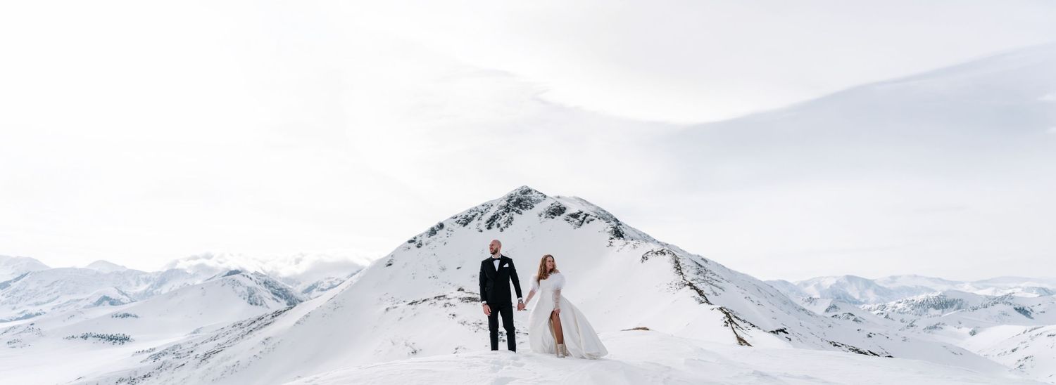 A couple in wedding attire poses against a stunning snowy mountain landscape on their winter wedding day.