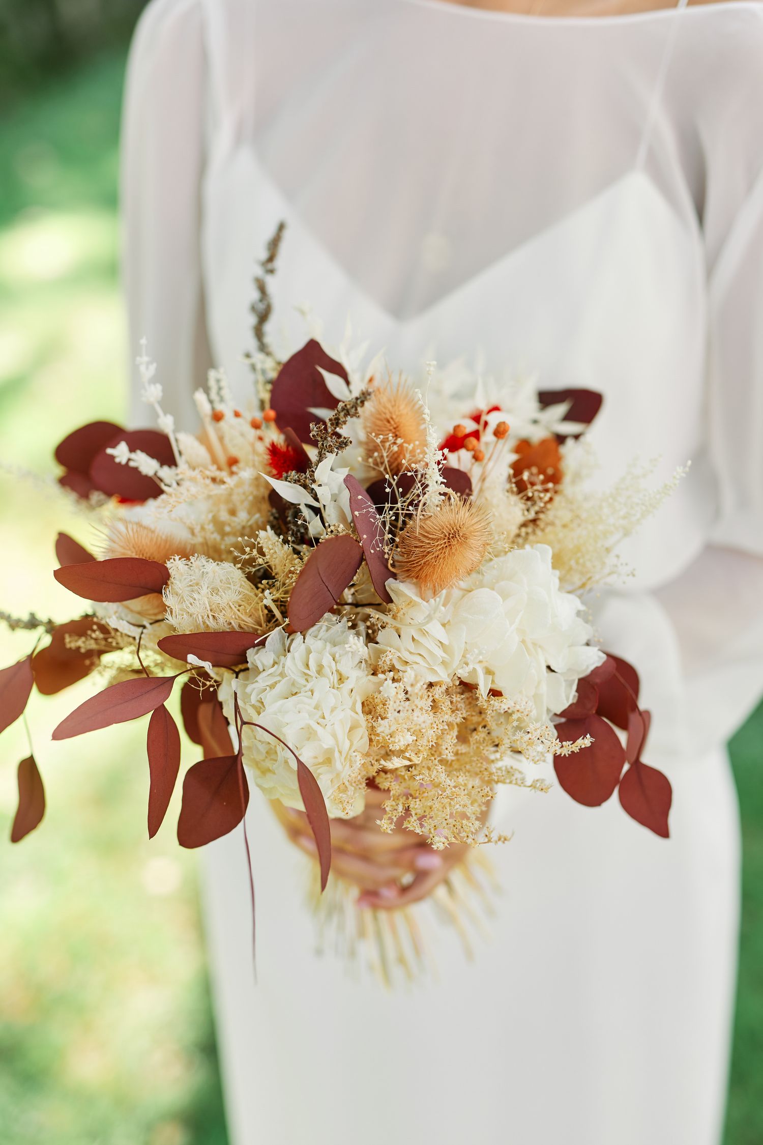 A delicate floral arrangement with burgundy leaves and white dried flowers against a soft white fabric background.