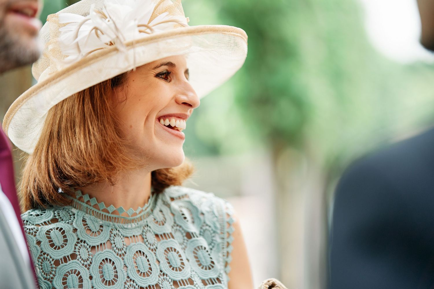 Elegant woman in a vintage-style dress and wide-brimmed hat smiles while standing outdoors in natural lighting.