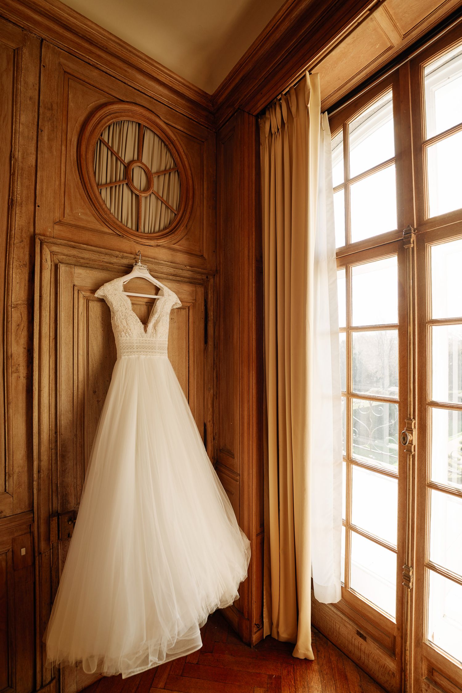 A white wedding dress hangs elegantly in a wooden-paneled room with sunlight streaming through tall windows.