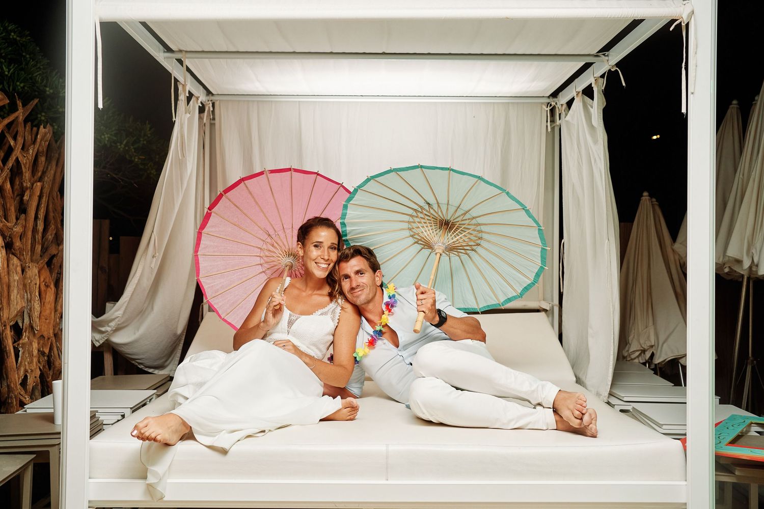 Romantic couple lounges on white daybed with pink and mint parasols in dreamy outdoor setting.
