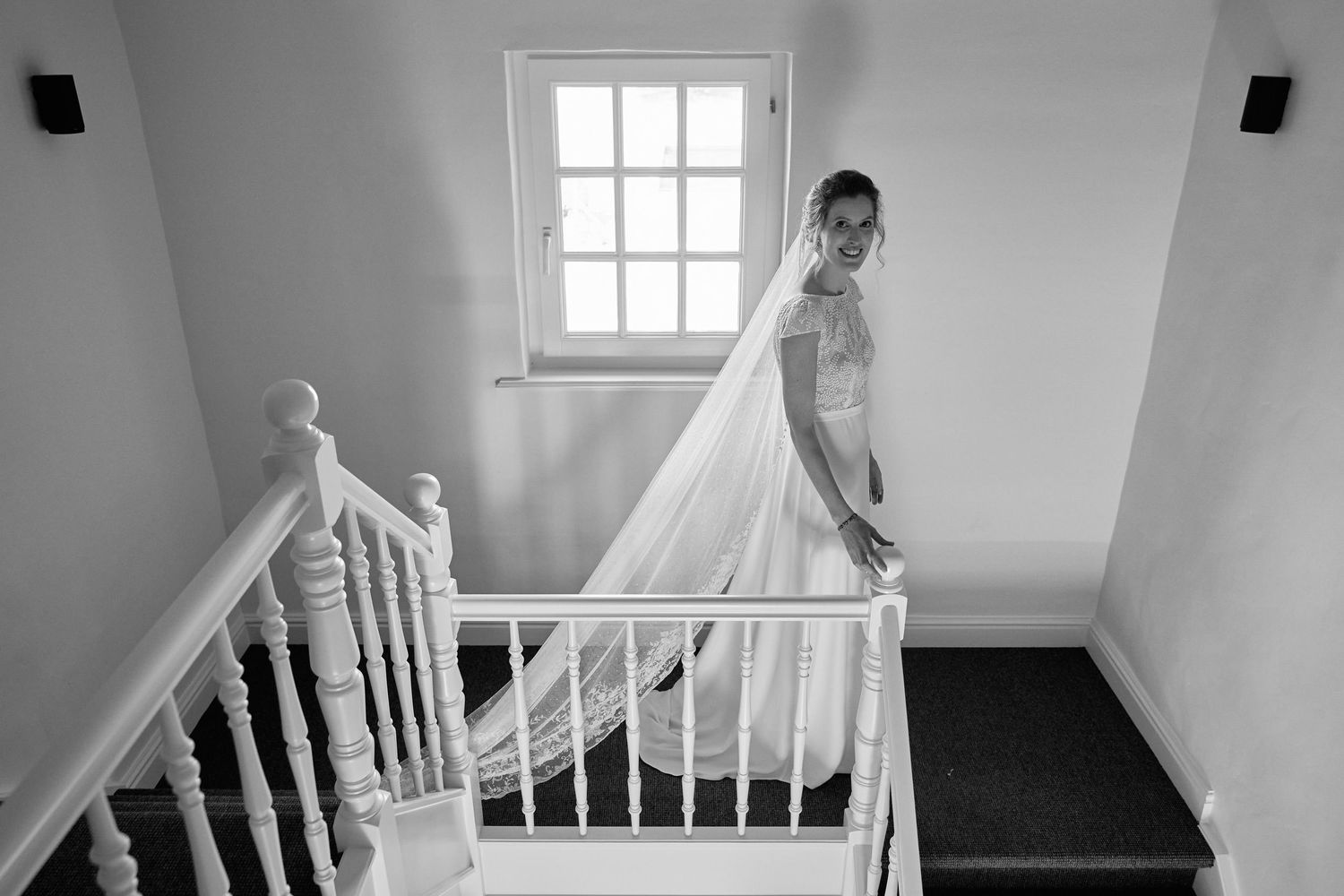 A bride in a flowing white veil stands at the top of a staircase in a minimalist white interior setting.