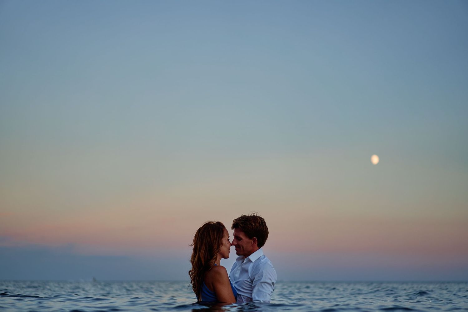 Couple embraces on beach at sunset with moon rising in pastel sky.