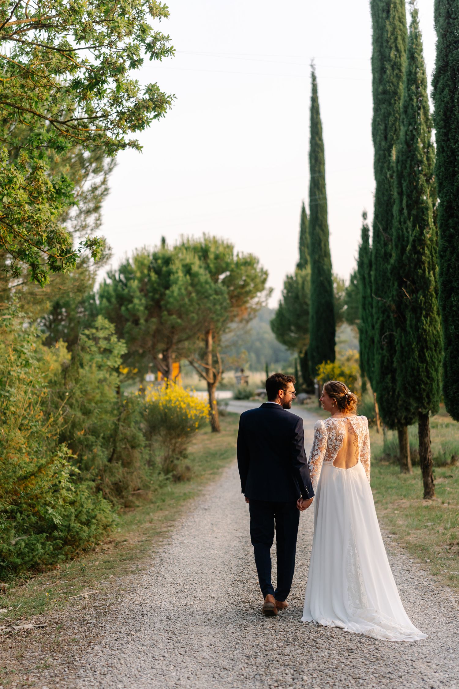 A couple in wedding attire walks down a cypress tree-lined path in the Italian countryside at sunset.