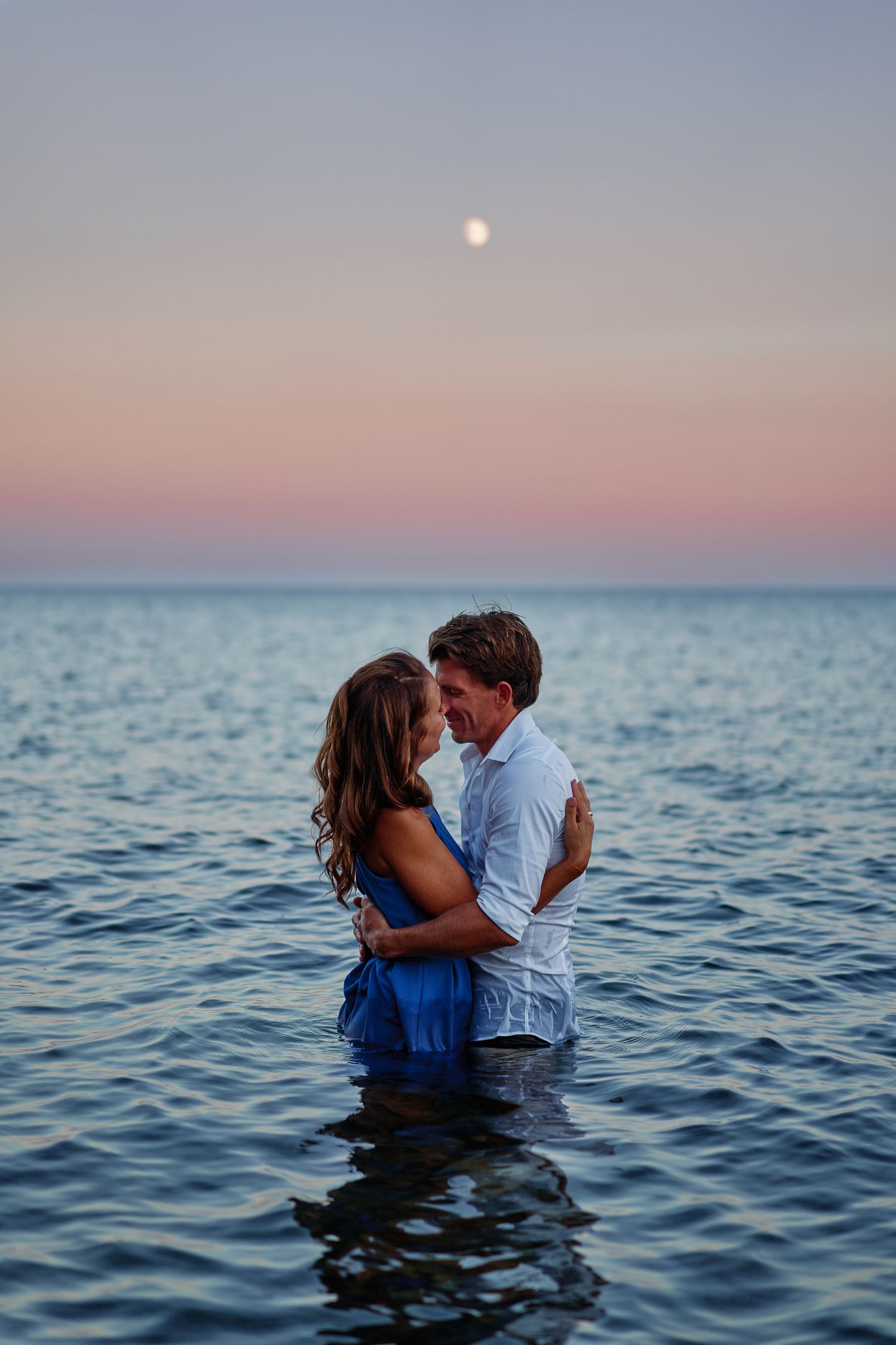 A romantic couple embraces while standing in calm ocean waters at sunset with a soft pink sky.