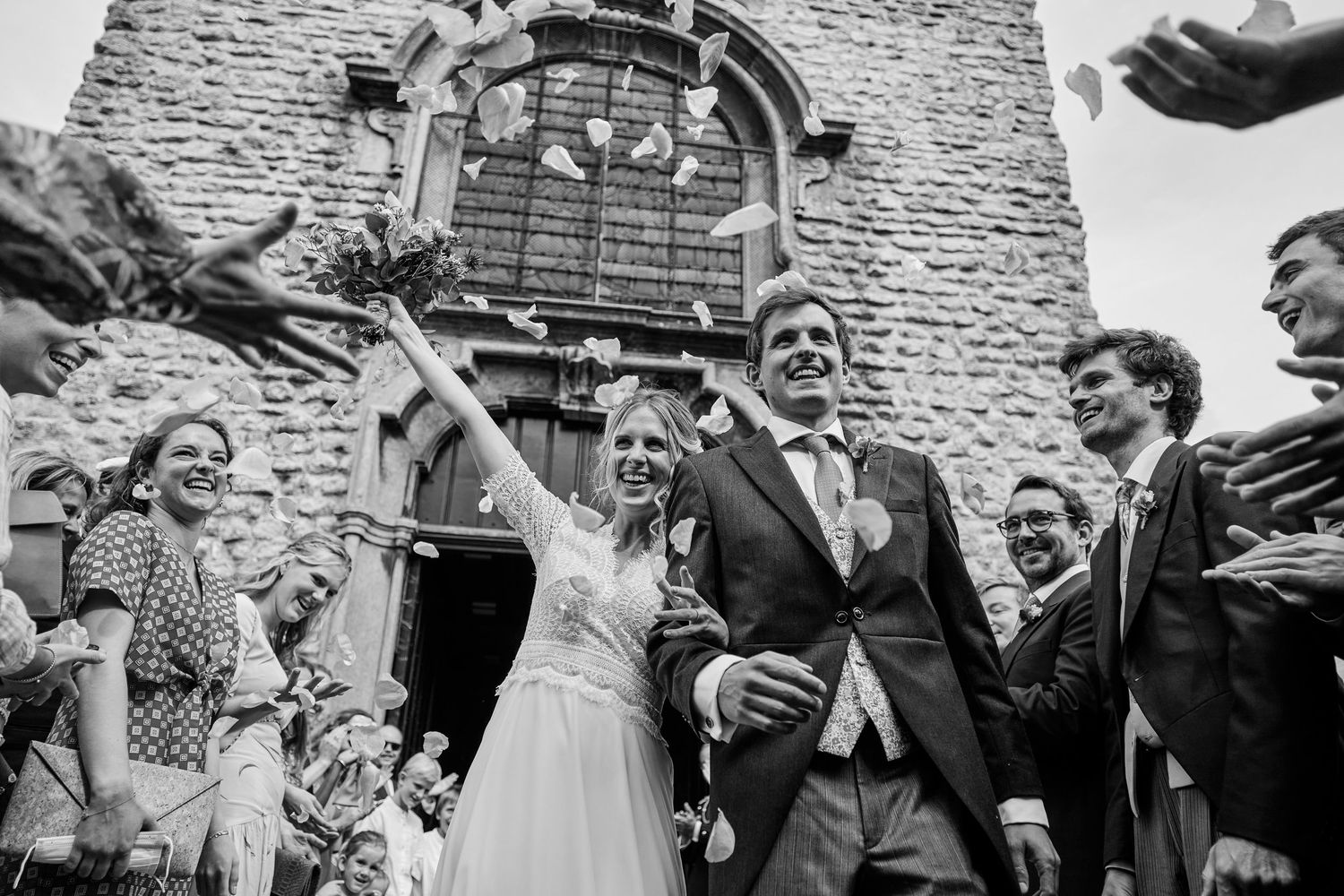 A joyful wedding celebration outside a stone church as newlyweds exit while guests throw confetti in black and white.