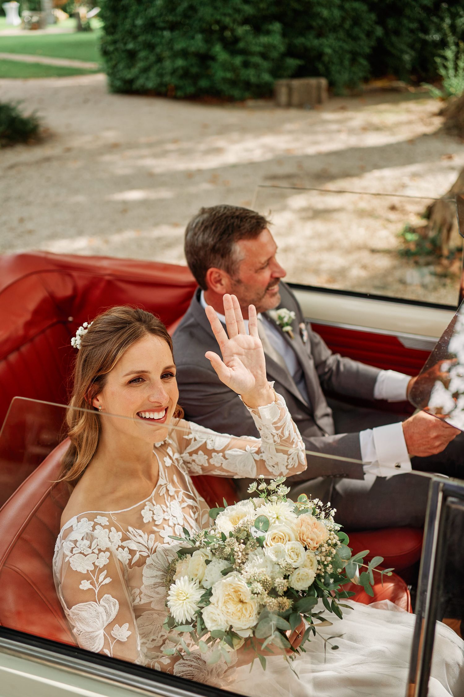 A happy couple waves from inside a vintage red convertible car during their wedding celebration.