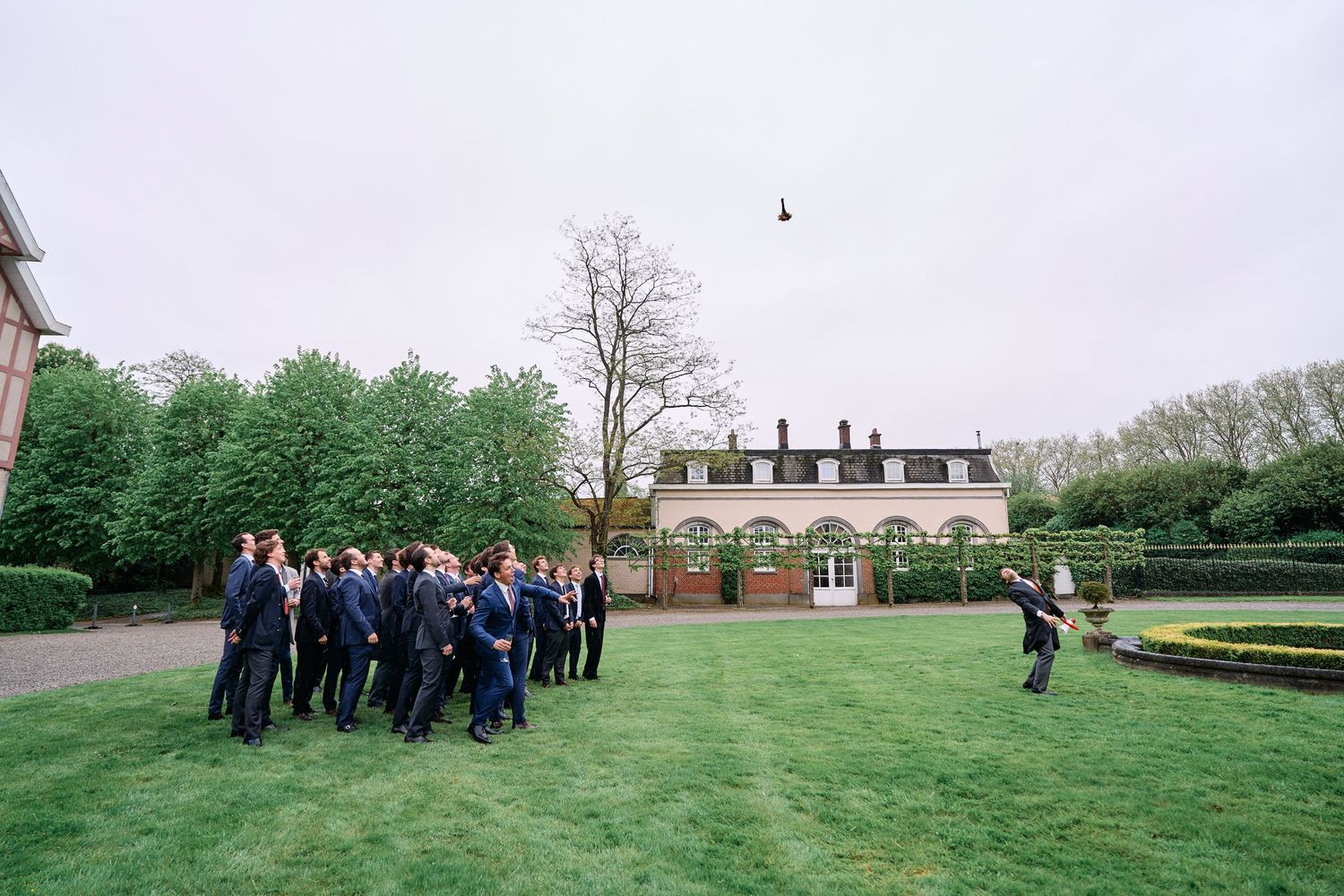 Wedding party gathers on a lush green lawn outside an elegant brick estate house during a spring celebration.