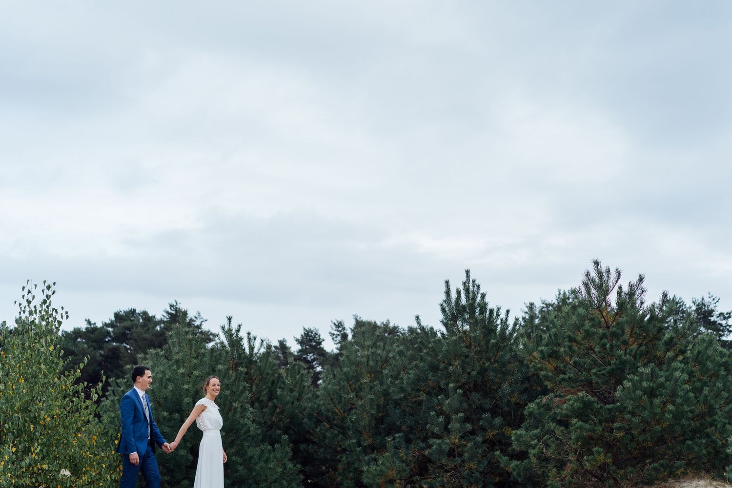 A couple walks together through a scenic outdoor landscape with evergreen trees and cloudy skies in the background.