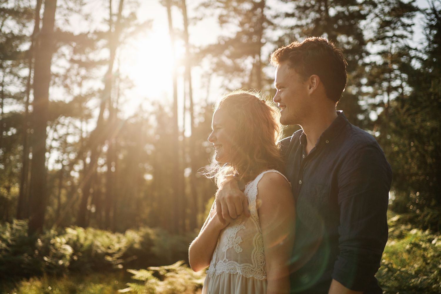 A couple embraces in a sunlit forest during golden hour as sunbeams filter through the pine trees.