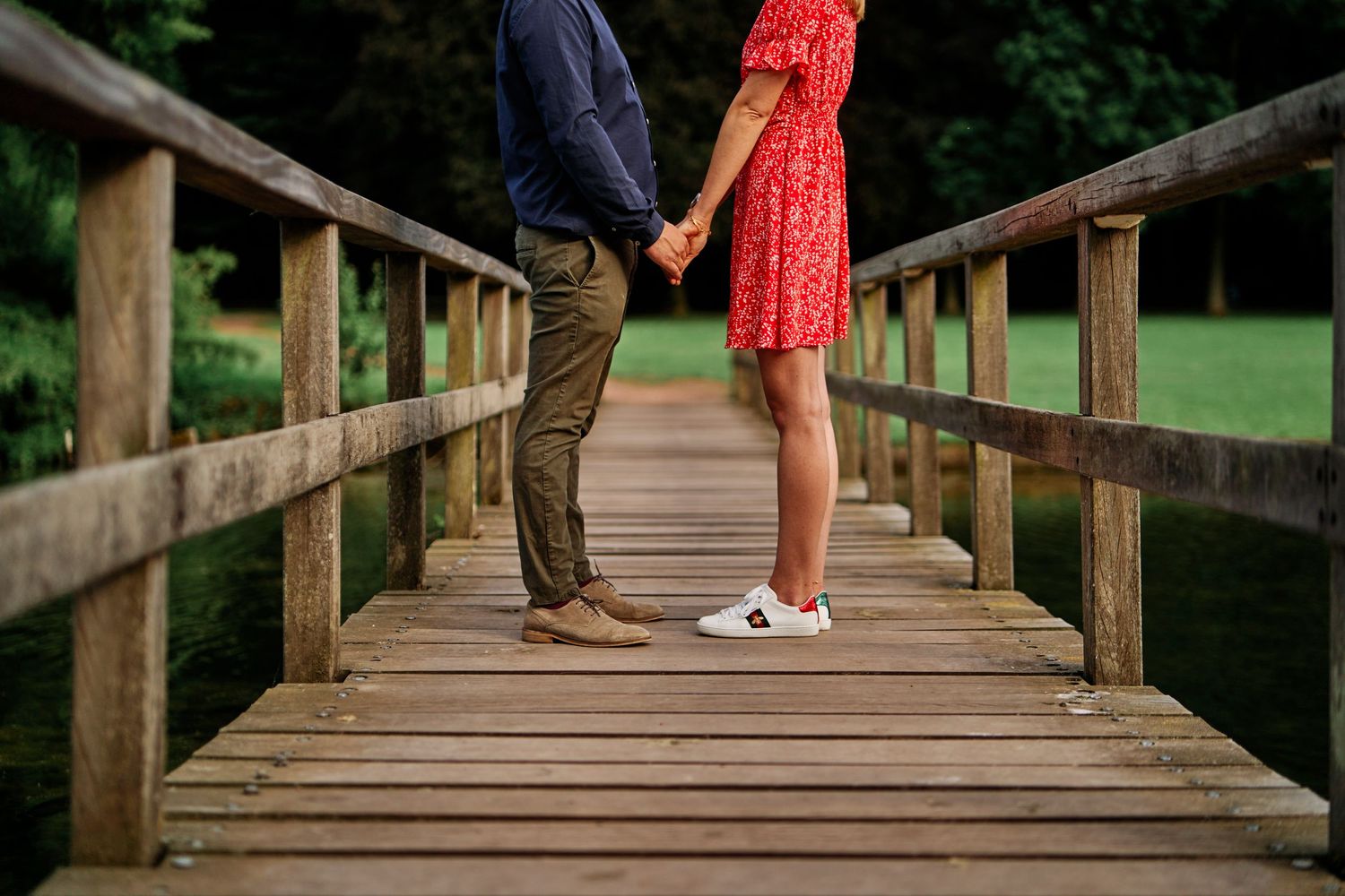 Couple stands together on a rustic wooden footbridge surrounded by lush green trees in a romantic outdoor setting.
