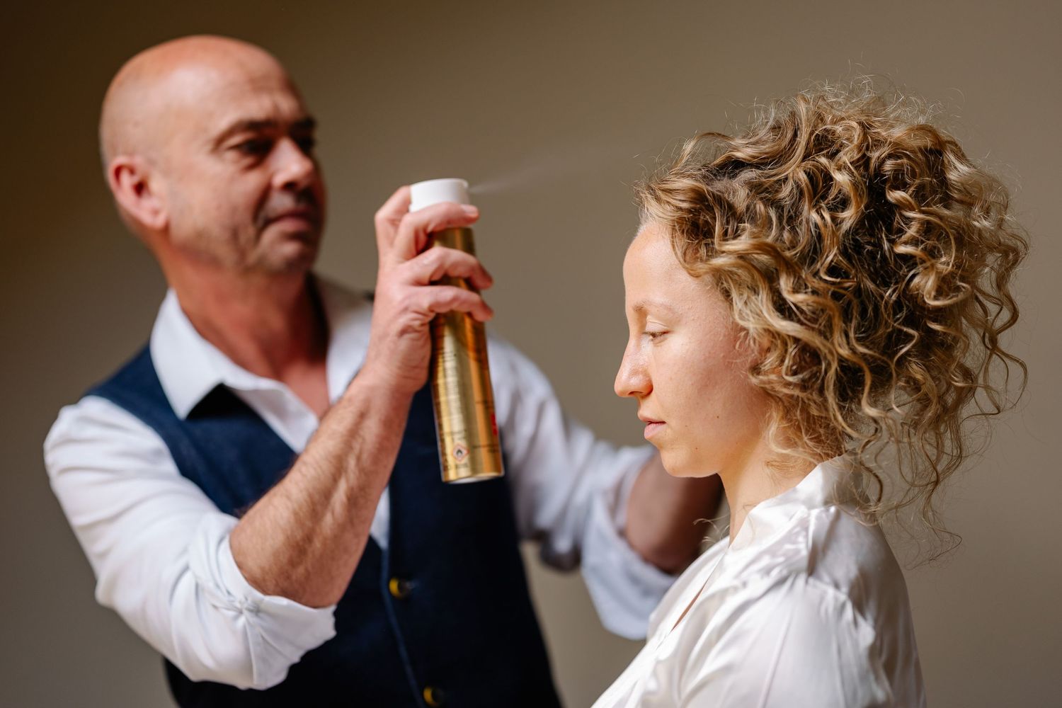 Professional hairstylist applies hairspray to style a bride's curly updo during wedding preparations.
