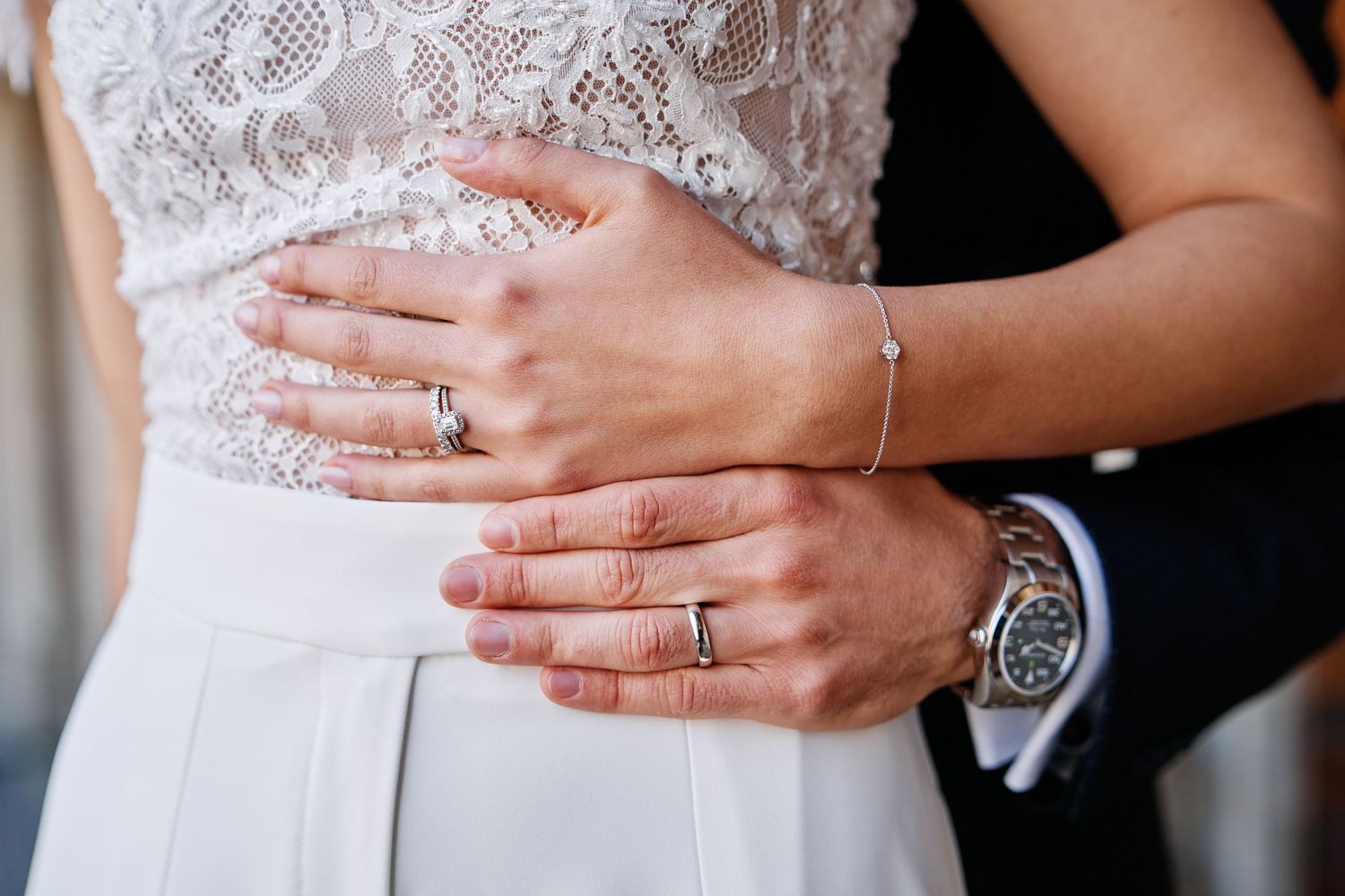 Close-up view of hands wearing wedding rings against a white lace wedding dress background.