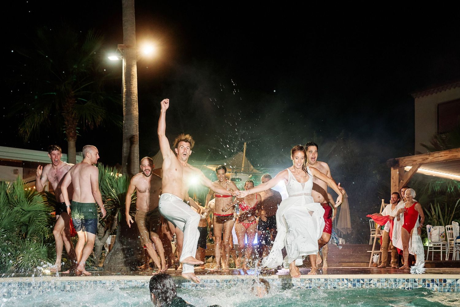 People dancing and having fun at an outdoor nighttime pool party with illuminated lights in the background.