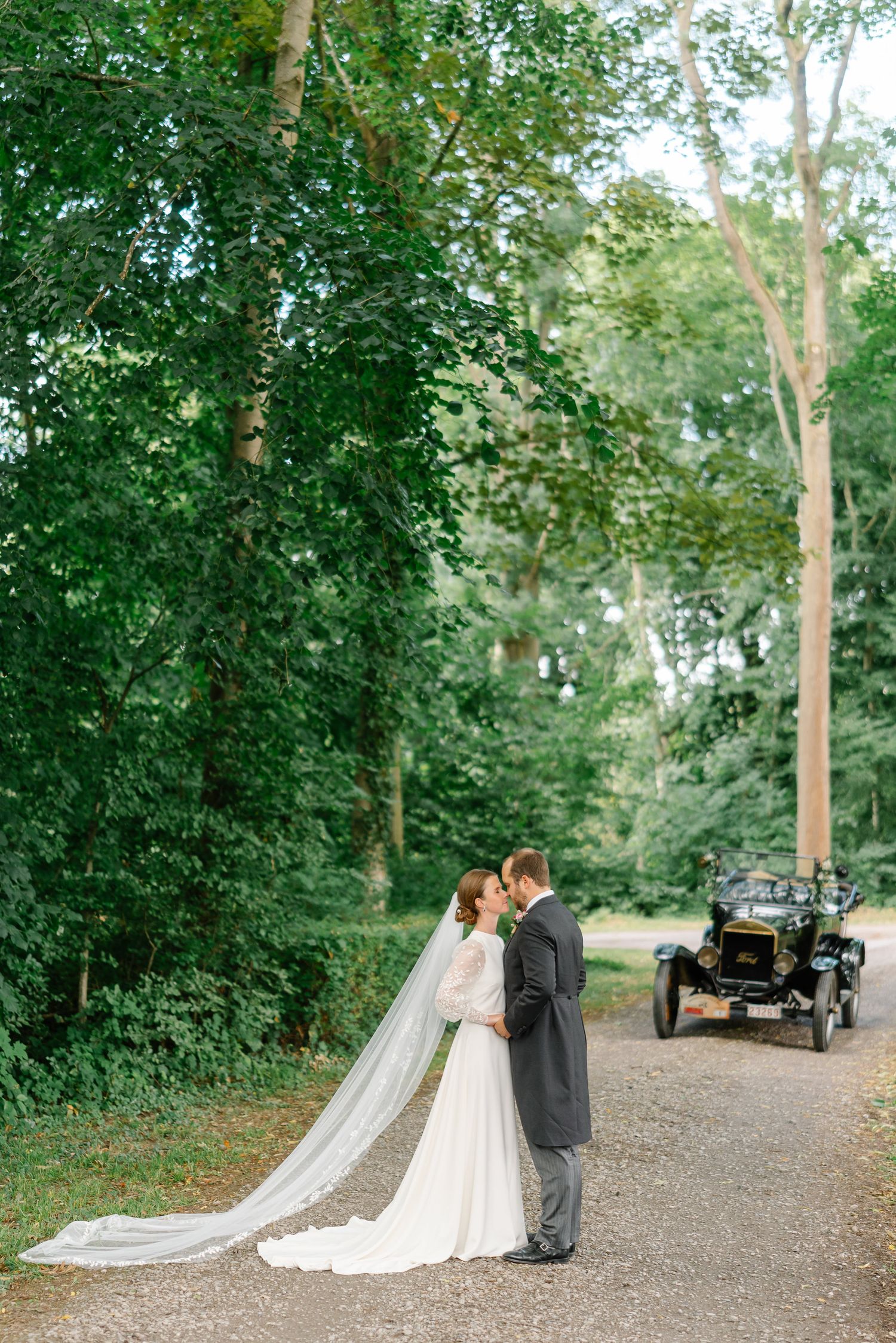 A romantic wedding couple embraces on a tree-lined gravel path with a vintage car visible in the background.