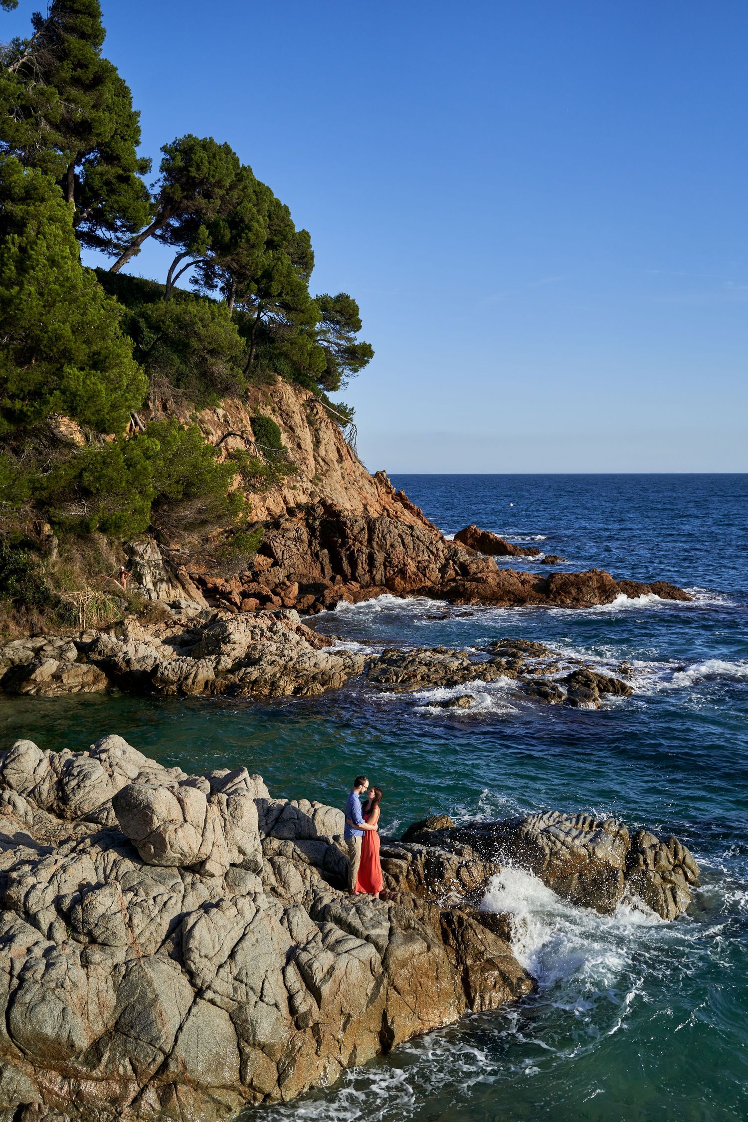 Rocky Mediterranean coastline with turquoise waters and pine trees along rugged cliffs on a sunny day.