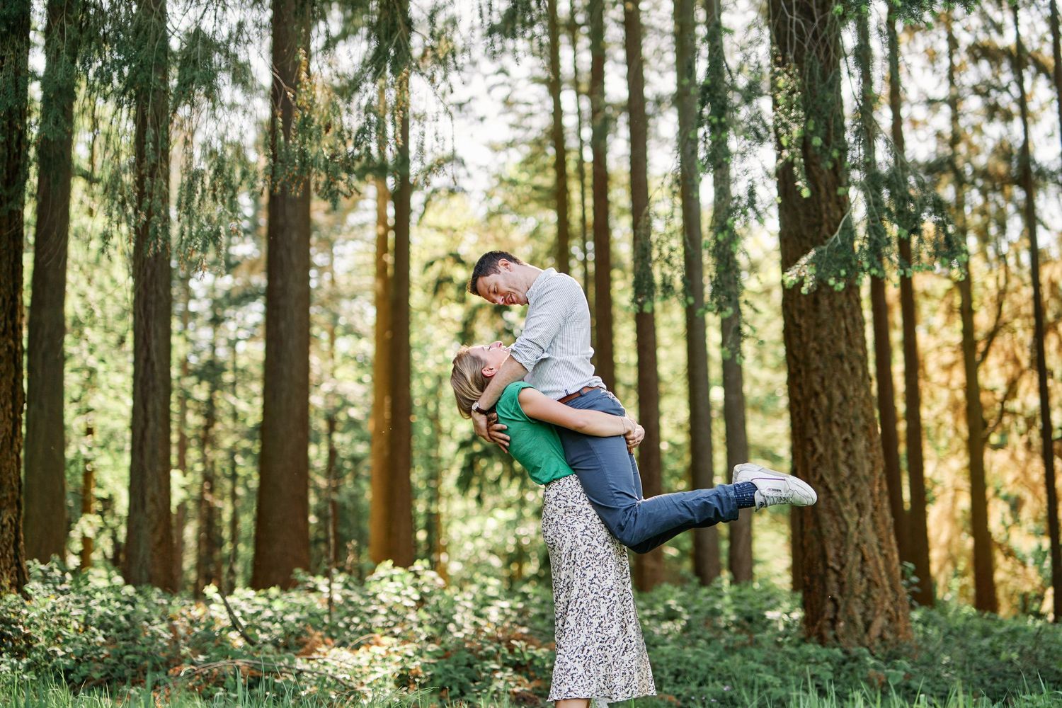 A couple embraces in a sunlit forest setting with tall pine trees creating a romantic woodland backdrop.