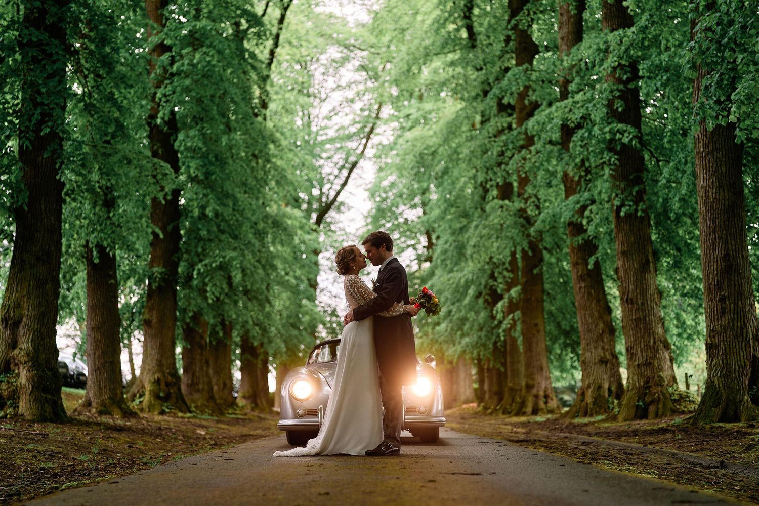 A couple embraces on a tree-lined road with vintage car headlights glowing behind them at dusk.