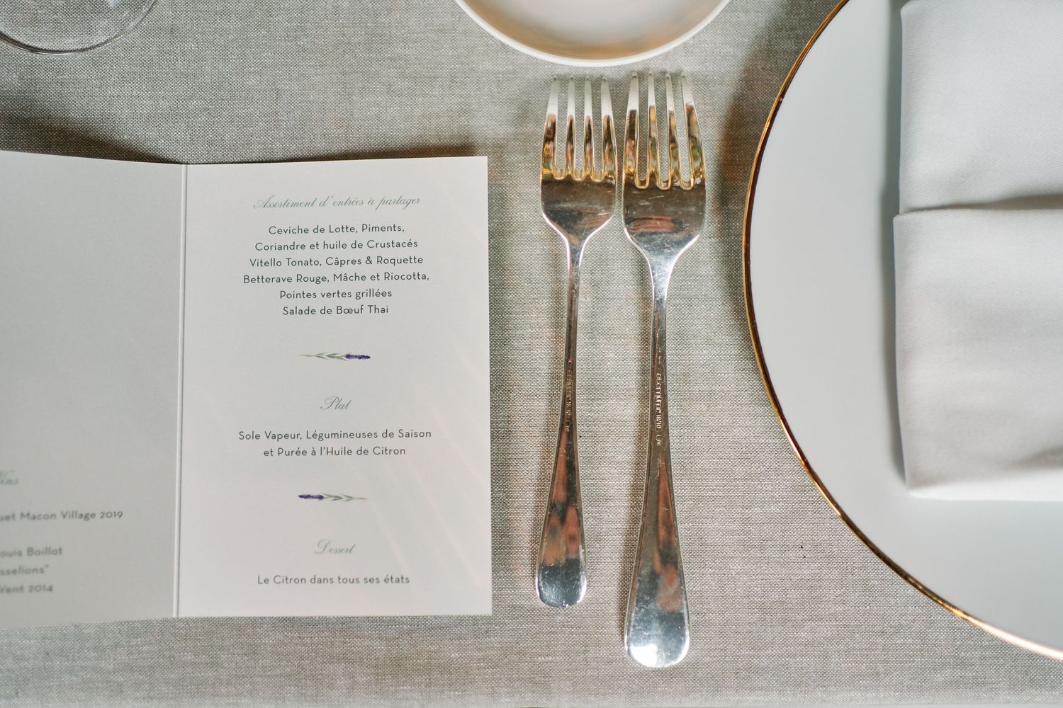 Elegant place setting with silver forks and menu card on linen tablecloth with white plate and gold trim.