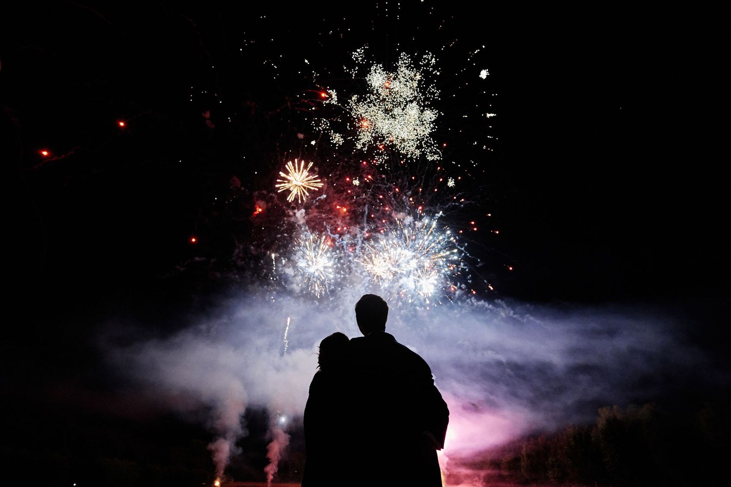 Silhouetted figure watches colorful fireworks display against dark night sky with bursts of red and white light.